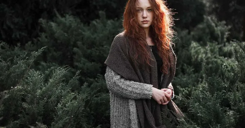 Jeune femme aux cheveux rouges dans une forêt, vêtue d'un cardigan gris et d'un manteau brun, les mains jointes